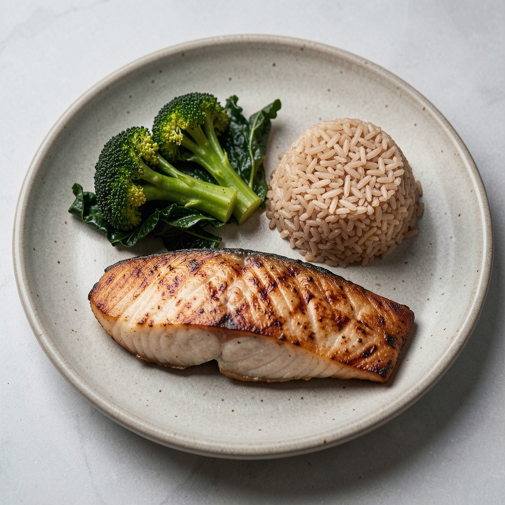 Stone-coloured plate showing balanced portion composition with a half serving of roasted broccoli and kale, palm-sized portion of grilled fish, and small mound of brown rice